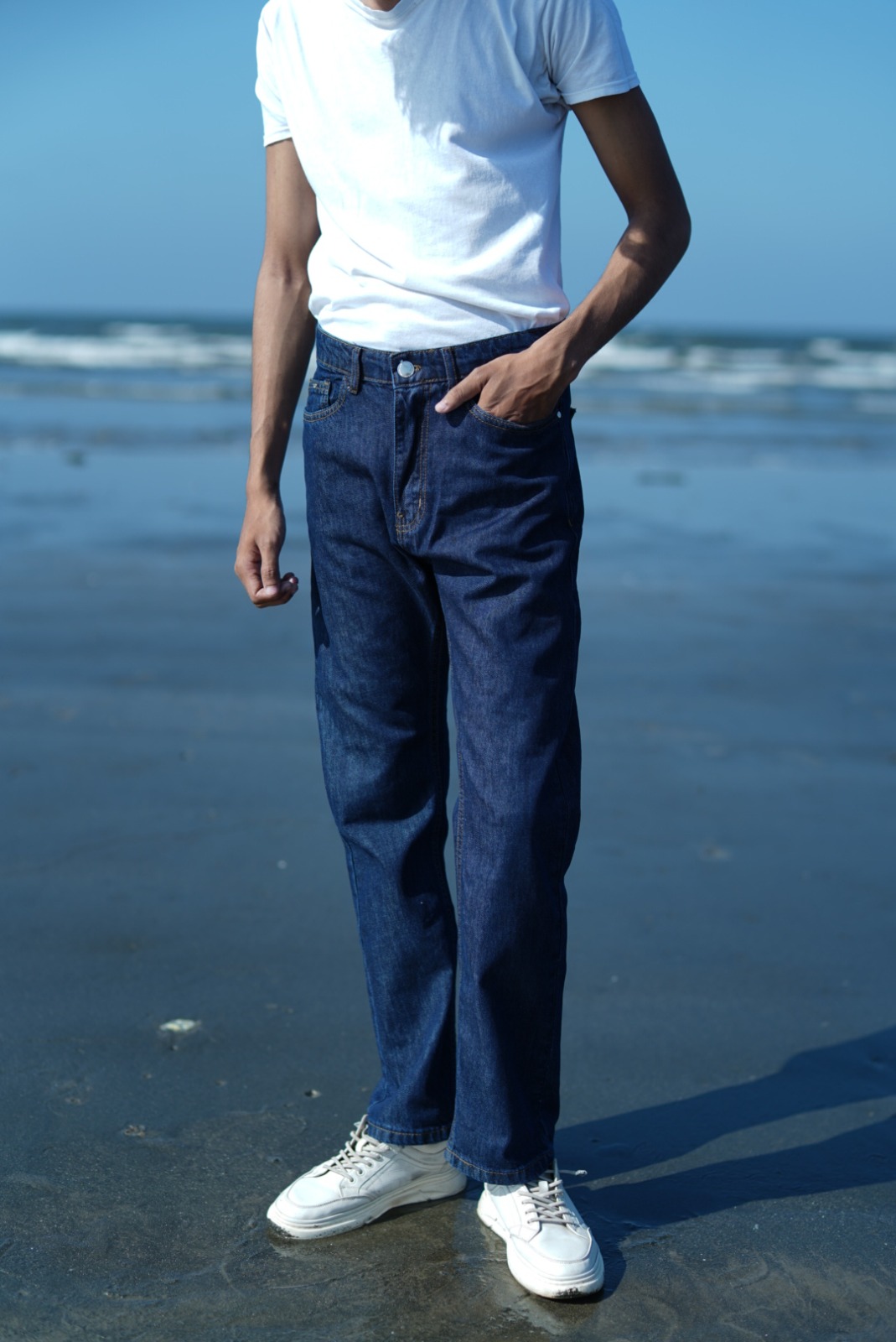 auto draft Men's dark blue straight fit denim jeans paired with a white t-shirt and white sneakers, photographed on a beach shoreline.